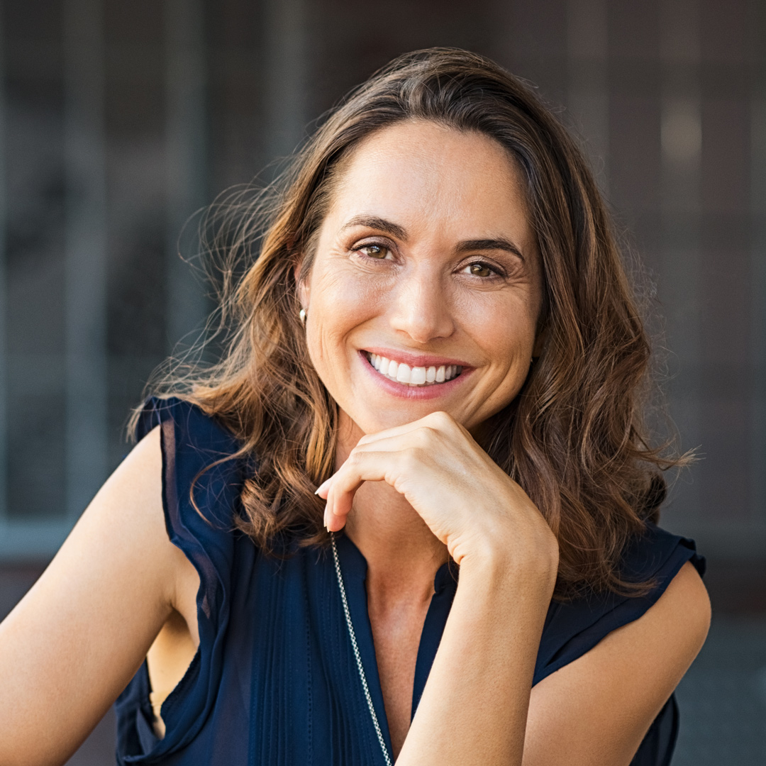 Begin-Your-Natural-Facial-Rejuvenation-Journey. A woman with wavy brown hair is seated with her elbow resting on a surface and her hand partially supporting her chin. She is wearing a sleeveless dark navy blouse with subtle pleated detailing and a long silver necklace. The background is softly out of focus, featuring a tiled or gridded surface.