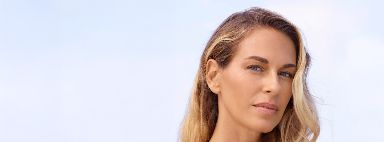 A close-up photograph of a woman with wavy blonde hair and blue eyes, looking towards the camera. She has a green, abstract-shaped facial mask applied to her cheeks, jaw, and the area around her mouth. The background is a soft, pale blue sky, and the image is in a wide banner format with ample copy space to the left.